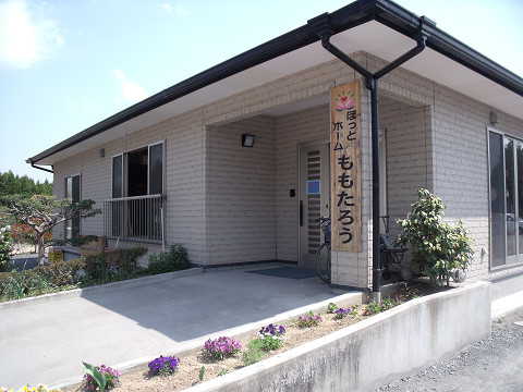 介護施設の建物外観。白い壁で平屋造り、カーポート付き。看板が設置されている。